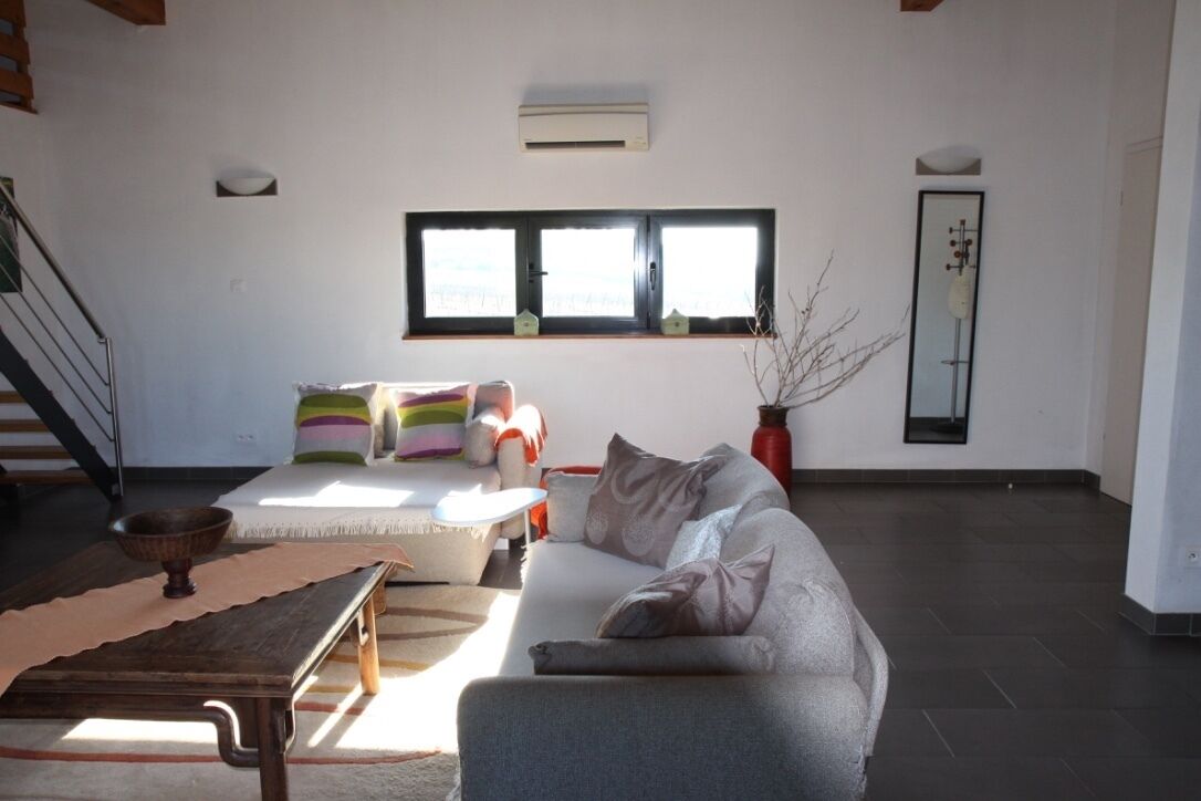 Ein Wohnzimmer mit grauem Sofa, bunten Kissen, einem Holztisch und einem Fenster mit Jalousien.