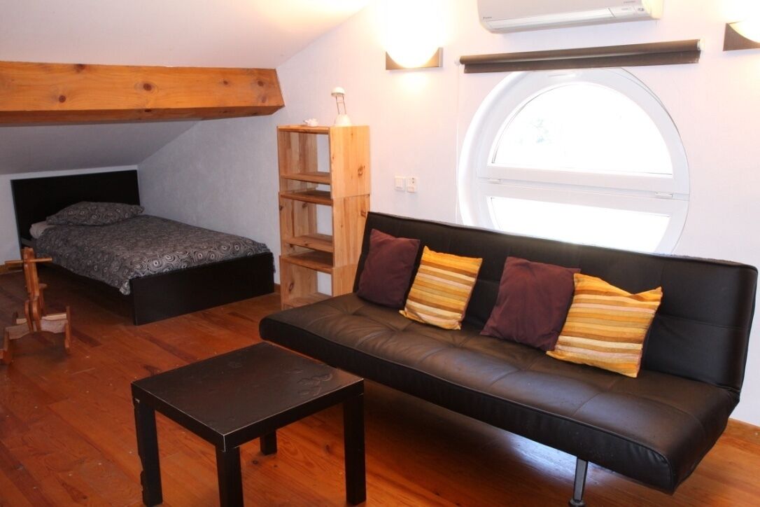 A small, cozy room with a loft bed, a black leather sofa with cushions, a wooden table, and a window with a view.