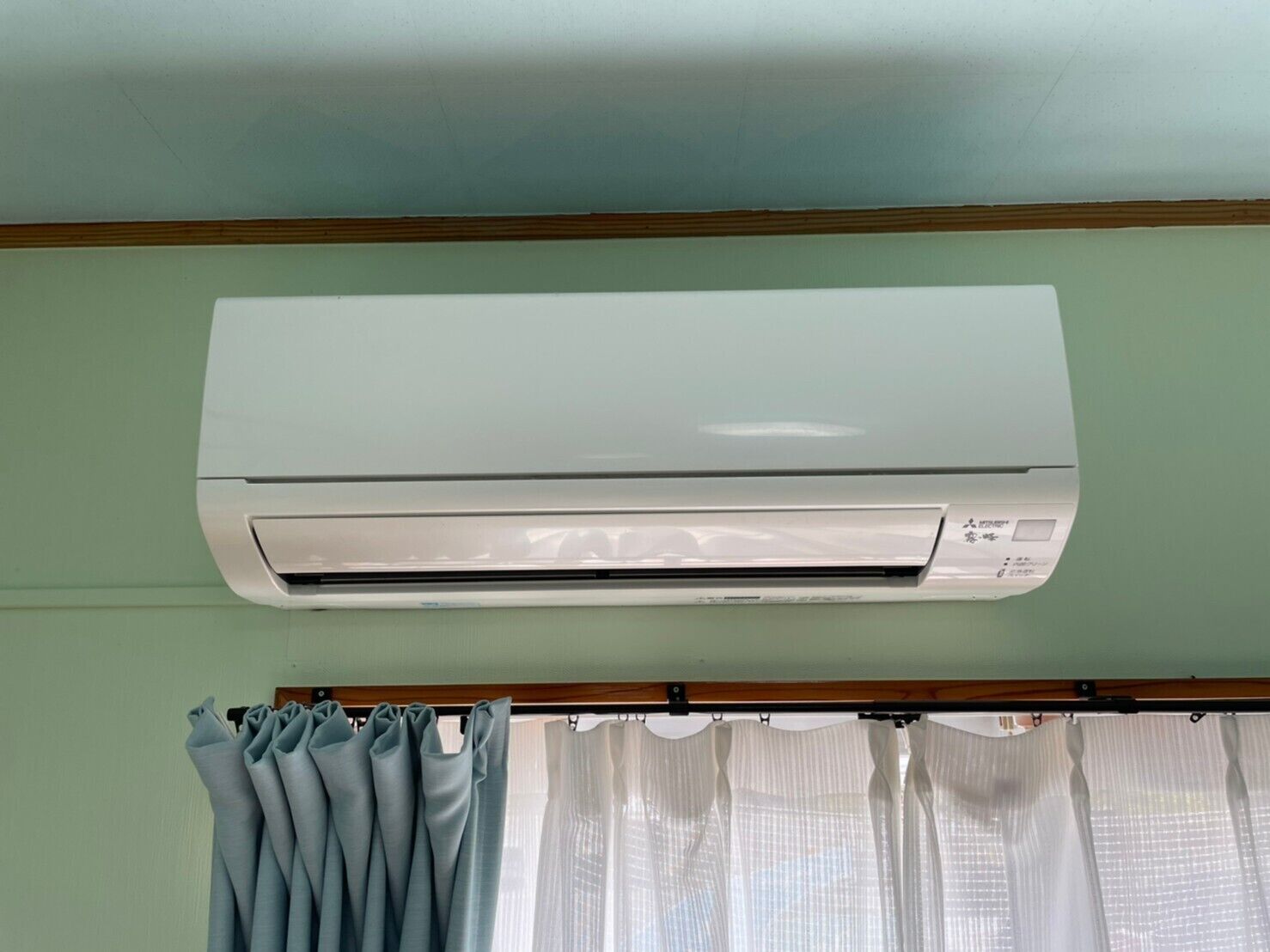 Una habitación con aire acondicionado blanco montado en la pared, techo de madera y ventana con cortinas azul claro.