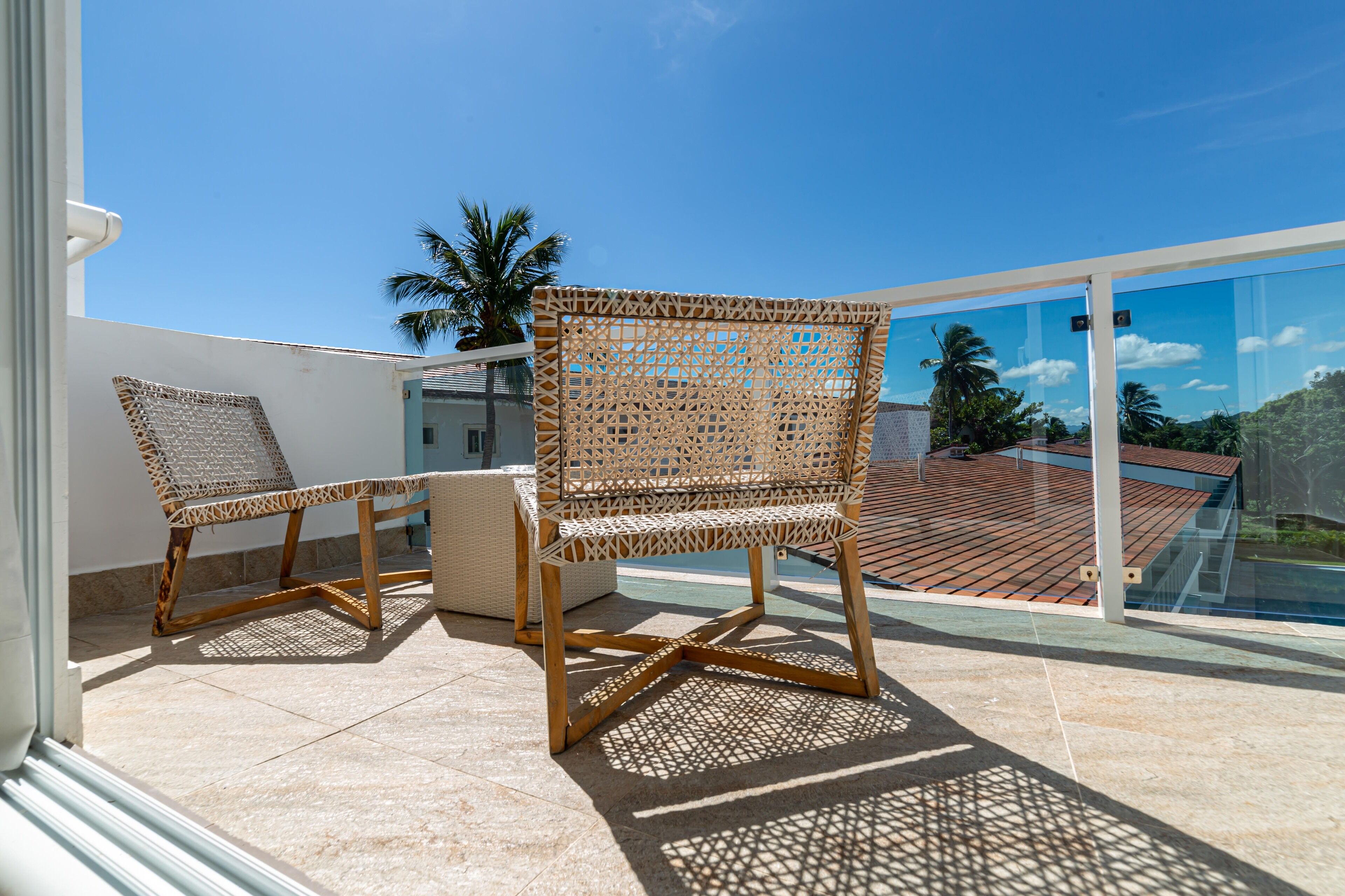 Un balcon avec deux fauteuils en osier et une petite table, donnant sur une terrasse carrelée et un ciel d’un bleu limpide.
