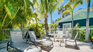 An outdoor patio with lounge chairs and a table, surrounded by palm trees and a white fence. - Blue Lagoon North by SeaBreeze Vacation (Anna Maria)