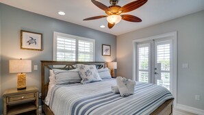 A bedroom with a bed, two nightstands, a ceiling fan, and two windows with shutters. - Blue Lagoon North by SeaBreeze Vacation (Anna Maria)