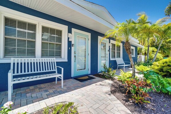 A house with a blue exterior, white trim, a front porch with a bench, and a small garden with palm trees and plants.