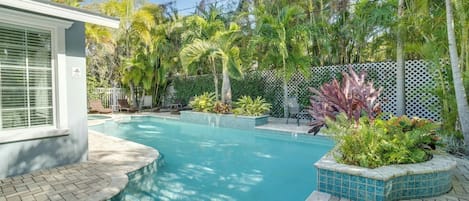 Une piscine dont le bord est recouvert de carrelage, entourée d’une clôture blanche, de palmiers et de diverses plantes.
