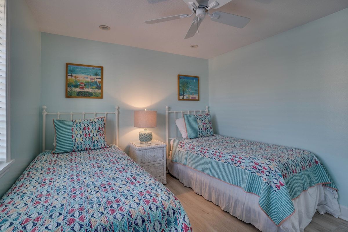 A bedroom with two beds, a ceiling fan, and two wall-mounted paintings.
