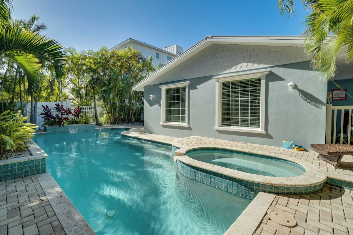 A swimming pool with a hot tub, surrounded by palm trees and a house with a light blue exterior.