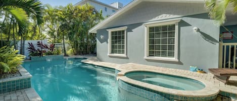 Una piscina con hidromasaje, rodeada de palmeras y una casa con fachada azul claro.