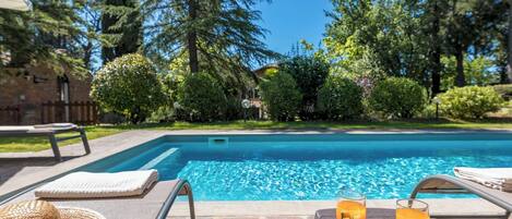 Area piscina con poltrone prendisole, un tavolo con frutta e bevande, e vista su una piscina circondata da alberi e un edificio.