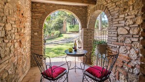 A stone-walled patio with two metal chairs and a table, featuring a view of a garden through an archway.
