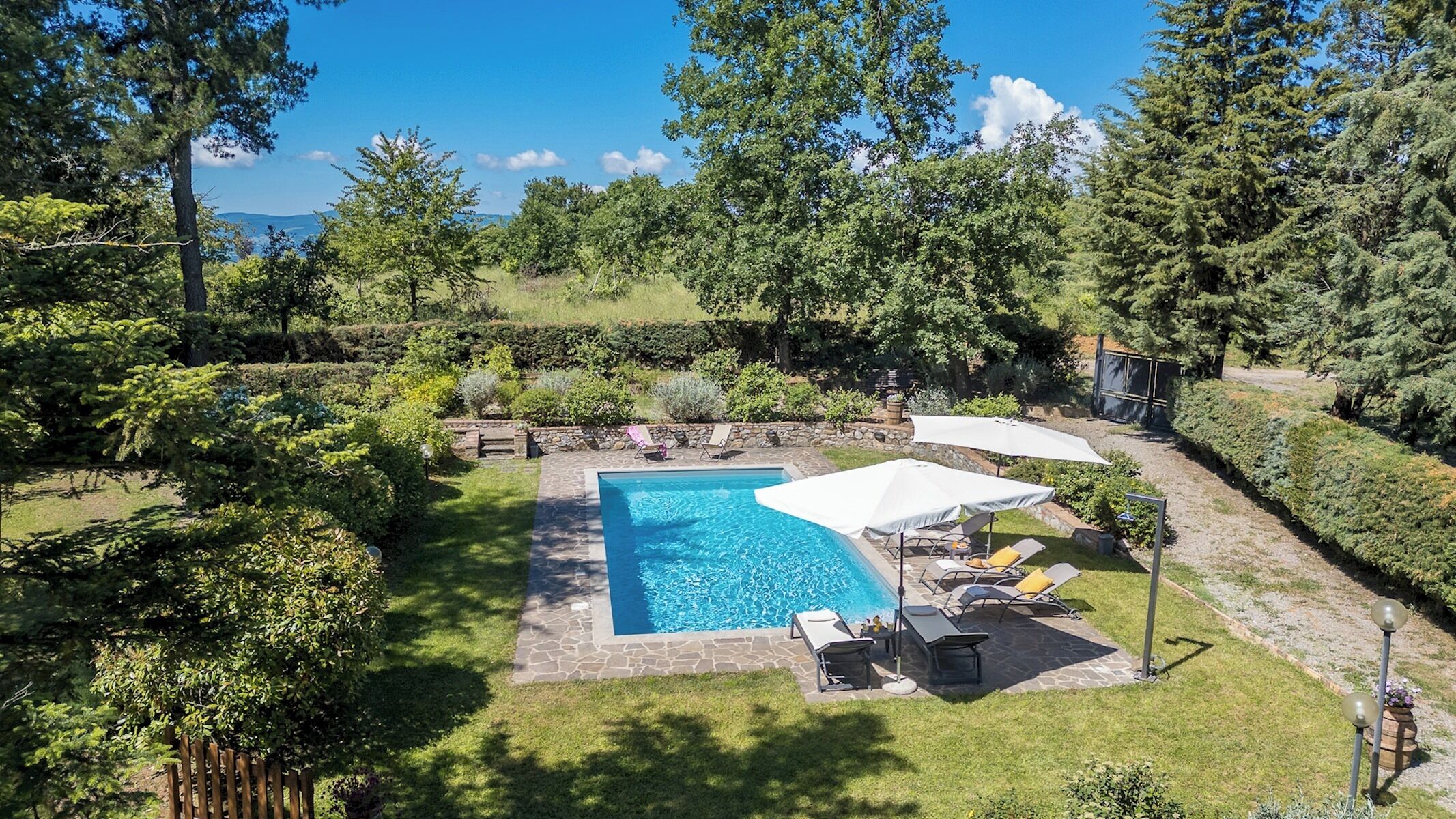 A rectangular swimming pool with a stone border, surrounded by a grassy area and trees. There are lounge chairs and umbrellas under a shaded area.