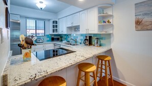 Una cocina moderna con armarios blancos, encimera de granito y estufa integrada. Hay dos taburetes de madera y una ventana con persianas.