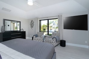 A bedroom with a bed, a dresser, a TV, a window with a view of a pool, and patterned chairs. - Tropical Waterfront Villa with Heated Pool & Spa (Punta Gorda)