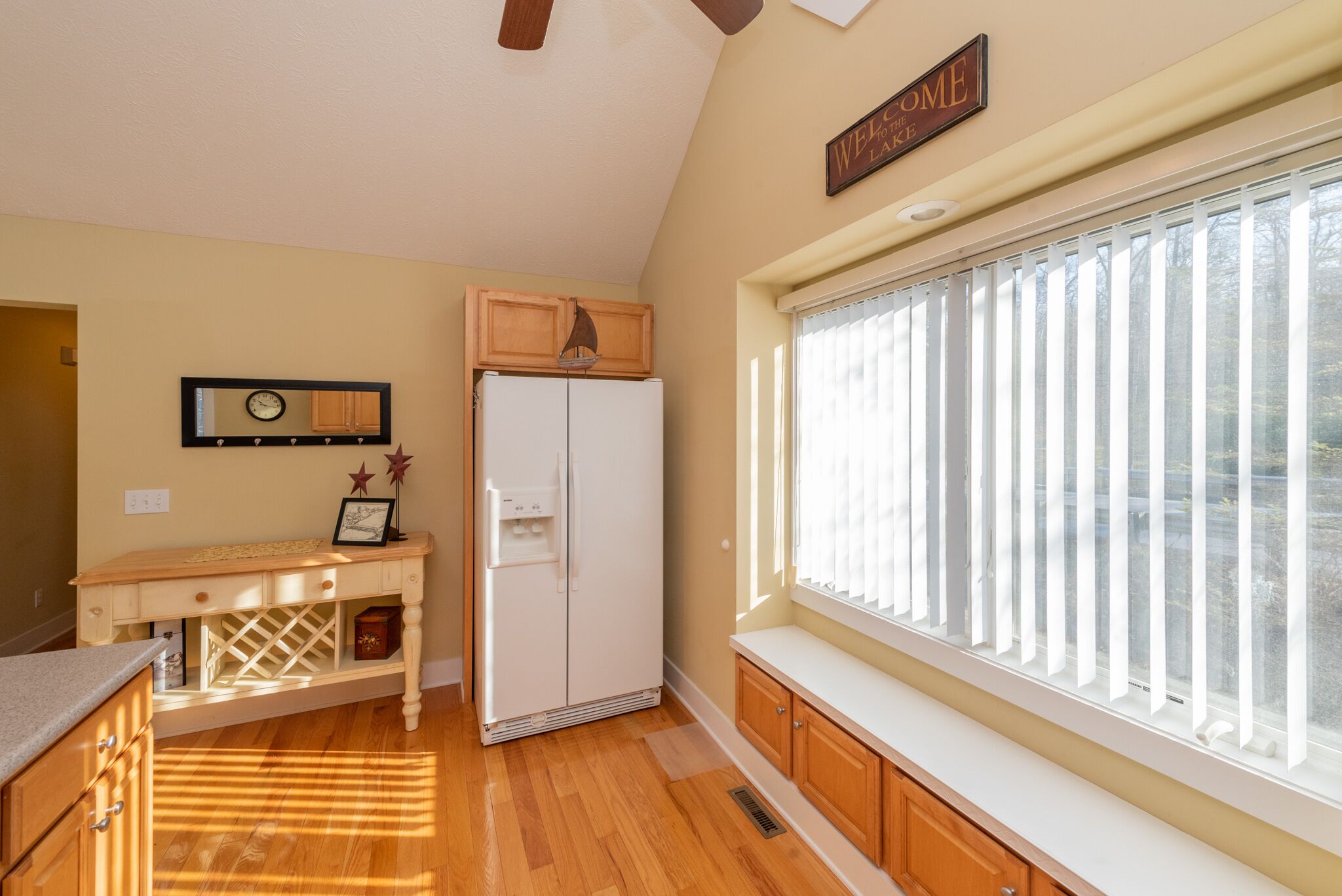 A kitchen with a refrigerator, wooden cabinets, a window with blinds, a clock on the wall, and a 'Welcome' sign.