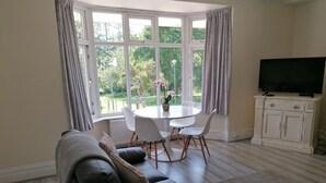 A living room with a black leather sofa, a white round dining table with chairs, a television on a white cabinet, and a view of greenery through large windows.