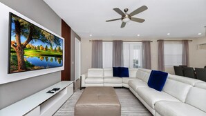 Una sala de estar moderna con un televisor de pantalla plana grande, un sofá seccional blanco, un reposapiés beige y un ventilador de techo.