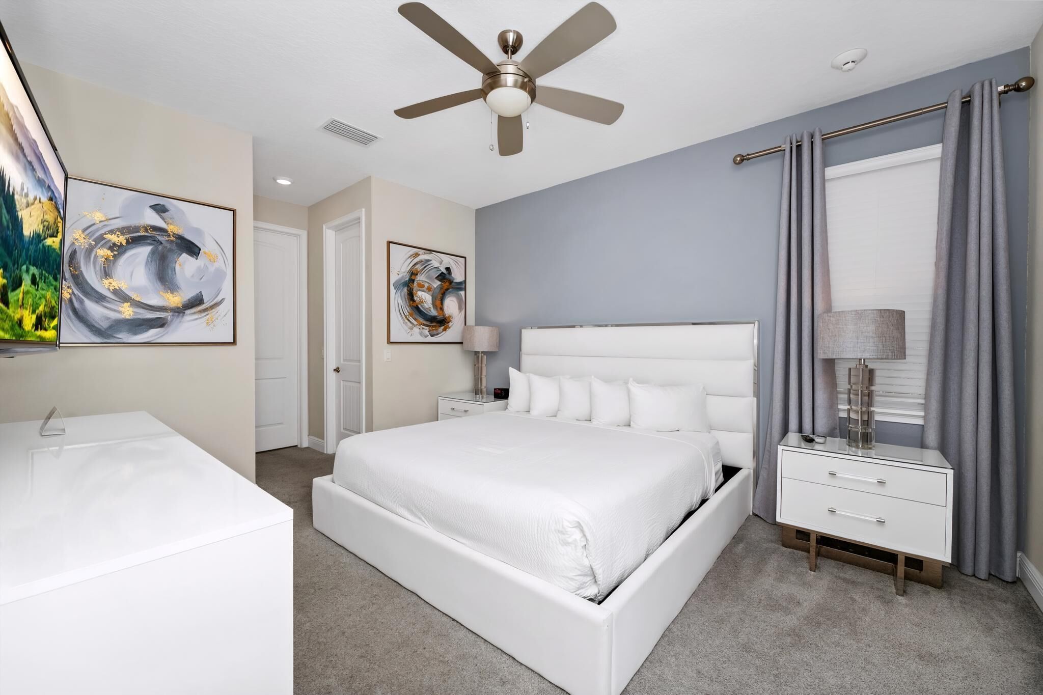 A modern bedroom with a large bed, a bedside table, a nightstand, a ceiling fan, and artwork on the walls.