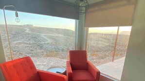 Ein Zimmer mit zwei roten Stühlen, einem kleinen Tisch, einer Pendelleuchte und großen Fenstern mit Blick auf eine hügelige Landschaft.