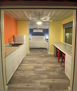 A narrow hallway with a kitchen on one side, a bed in the center, and a window on the other.