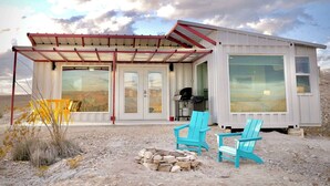 A modern, single-story building with large windows, a covered patio area, and two outdoor chairs. - Fossil Knob Ridge -Mariscal: Ultra Modern Container Home (Terlingua)