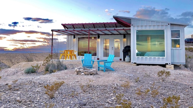 A small, single-story building with a covered patio area, outdoor seating, and a large window. - Fossil Knob Ridge -Mariscal: Ultra Modern Container Home (Terlingua)