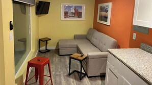 A compact living space with a sofa, a small table, a red stool, a ceiling fan, and two framed pictures on the walls. - Fossil Knob Ridge -Mariscal: Ultra Modern Container Home (Terlingua)