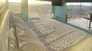 A bedroom with a large bed, a ceiling fan, a window with a view of a landscape, and a small table with a candle. - Fossil Knob Ridge -Mariscal: Ultra Modern Container Home (Terlingua)