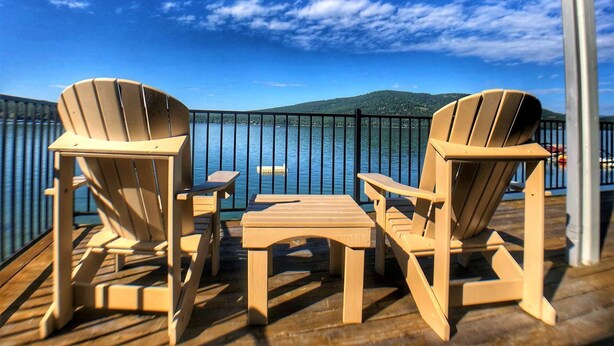 A deck with Adirondack chairs and a table, overlooking a lake with mountains in the background.