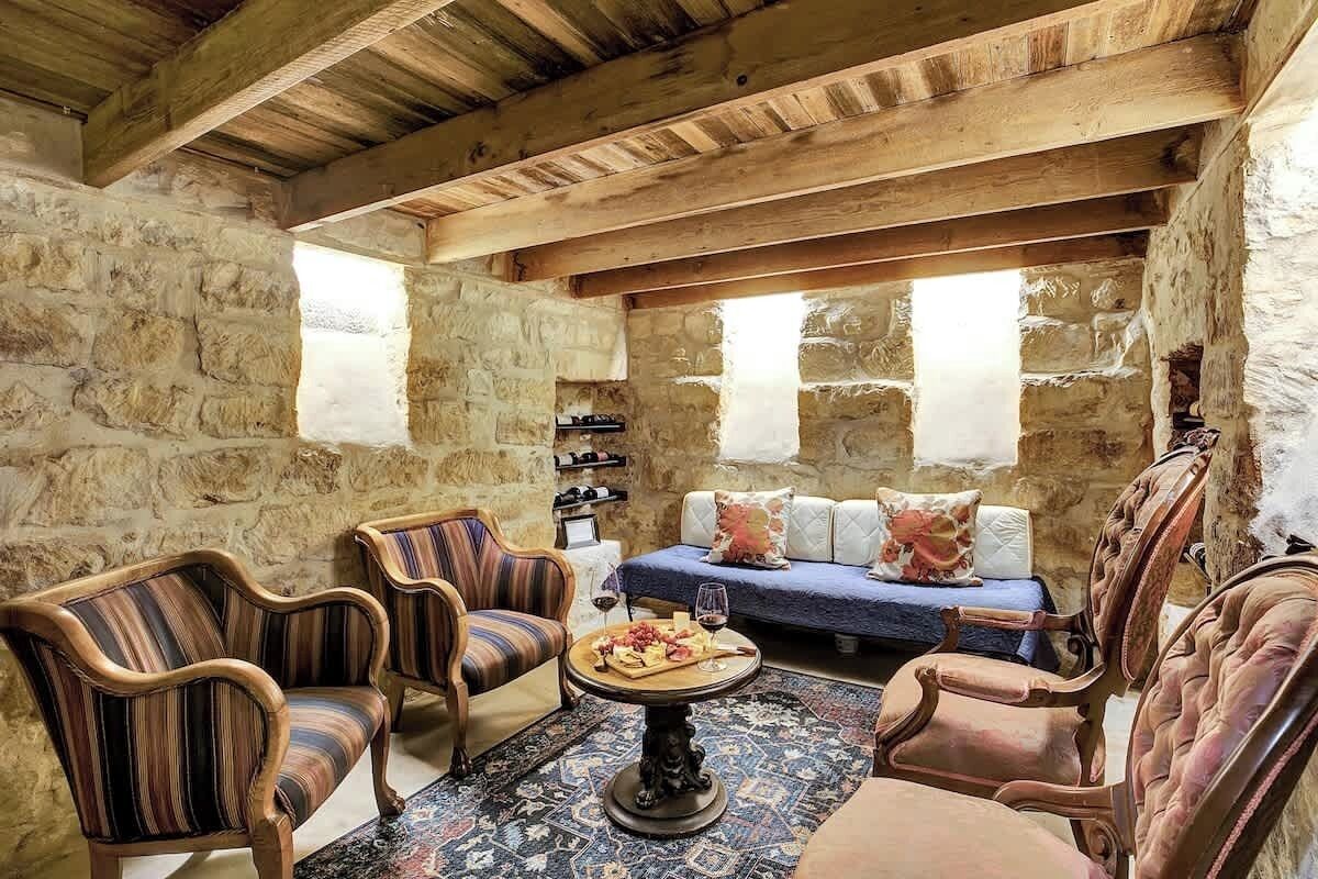 A rustic stone-walled room with wooden beams, a sofa, striped armchairs, and a small table with a glass of wine and cheese.