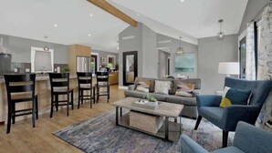 A spacious living area with a sofa, armchairs, and a coffee table. There is a kitchen area with modern appliances and a dining table with chairs. The room has a high ceiling with exposed beams and a large window providing natural light.