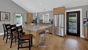 A modern kitchen with a central island, bar stools, wooden chairs, stainless steel appliances, and a view of greenery outside.