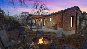A rustic outdoor patio with a fire pit, seating, and a covered area with a brick building in the background.