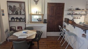 Een eethoek met een houten tafel, stoelen en een spiegel erboven. Er is een keuken met een spoelbak, planken en diverse keukenbenodigdheden.