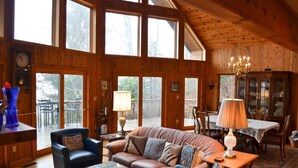 A spacious living room with a brown leather sofa, a blue armchair, and a wooden dining table with chairs. Large windows allow natural light to fill the room.