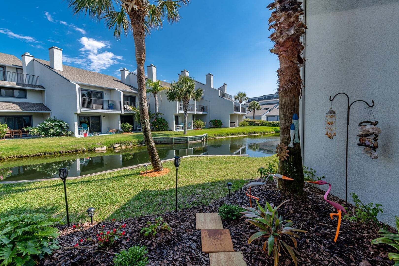 A residential area with a pond, palm trees, and a pathway leading to a building with balconies.