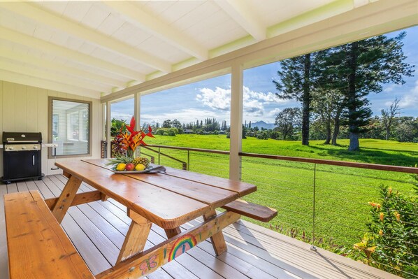 A wooden deck with a picnic table, a grill, and a view of a green field and trees.