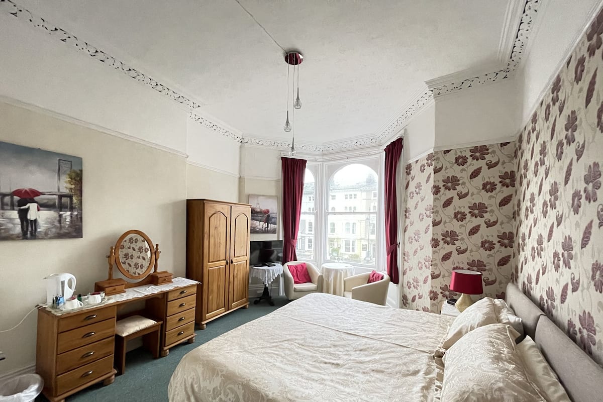 A bedroom with a bed, a desk, a wardrobe, and a window with a view of buildings.