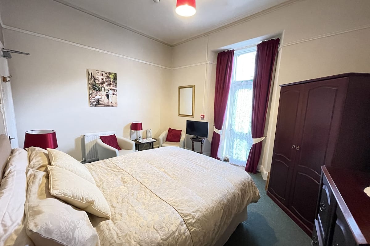 A hotel room with a large bed, two armchairs, a desk with a lamp, a television, and a wardrobe.