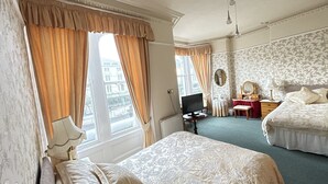 A hotel room with two beds, a desk with a computer, a chair, a small table with a lamp, and a view of the city through large windows. - White Court Llandudno (Llandudno)