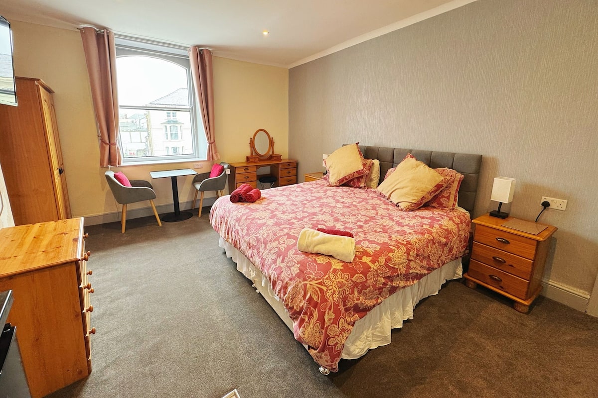 A bedroom with a bed, desk, chair, dresser, and a window with curtains.