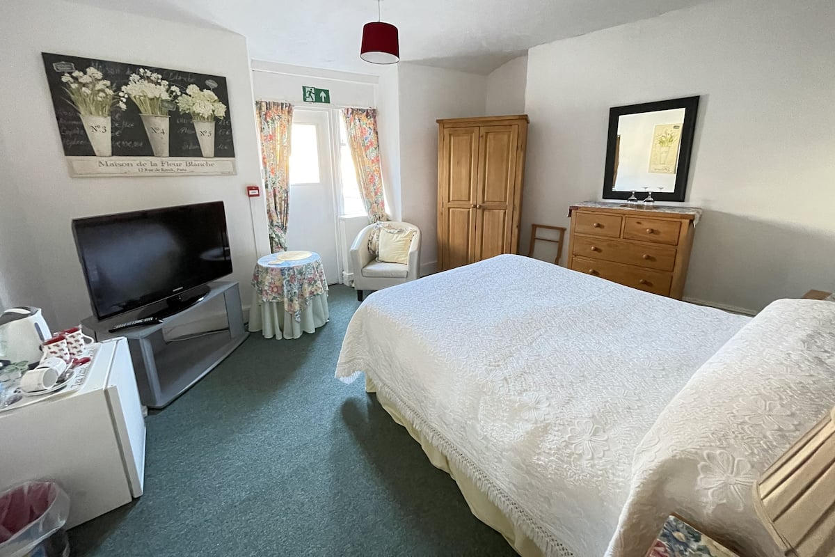 A hotel room with a bed, a TV, a wooden wardrobe, a small table with a floral tablecloth, and a chair.