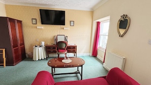 A room with a red sofa, a wooden table with a vase, a TV on the wall, a desk with a chair, a wardrobe, and a window with curtains. - White Court Llandudno (Llandudno)