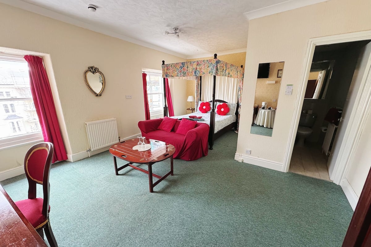 A hotel room with a bed, a red sofa, a wooden table, and a chair.