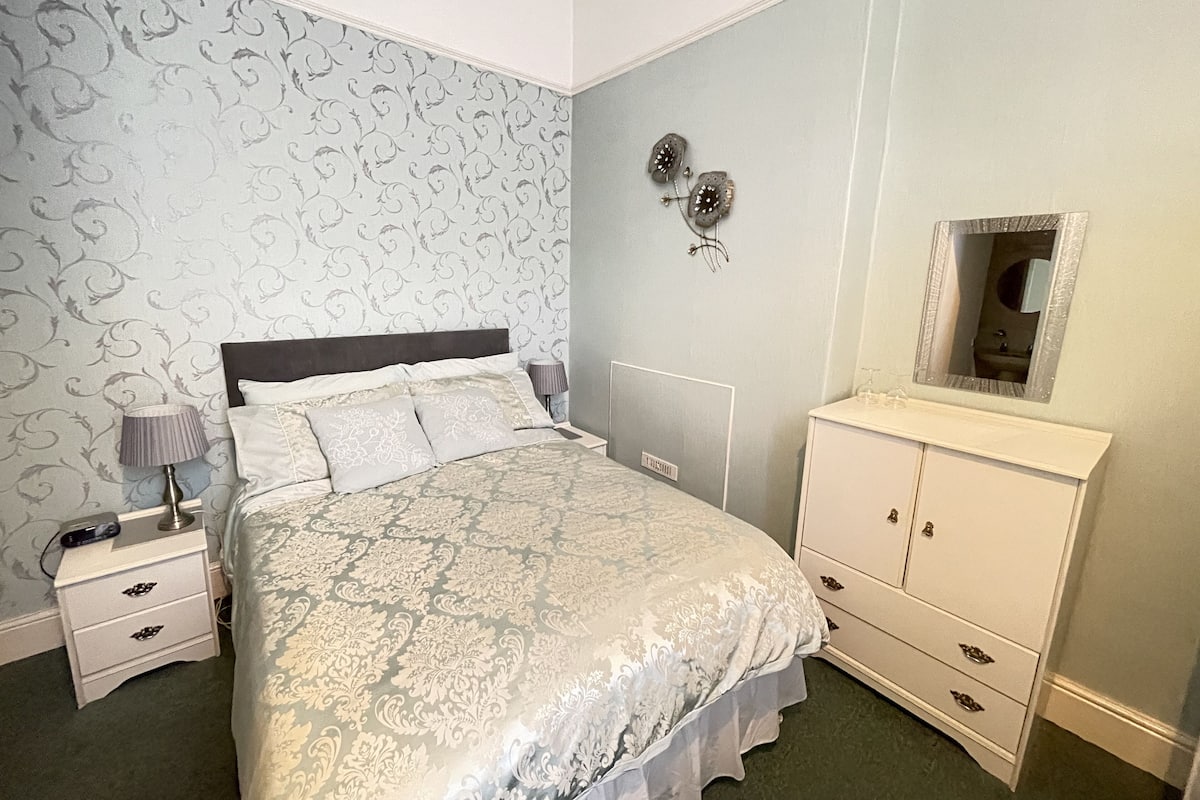 A neatly arranged bedroom with a bed, bedside table, lamp, dresser, and wall-mounted mirror.