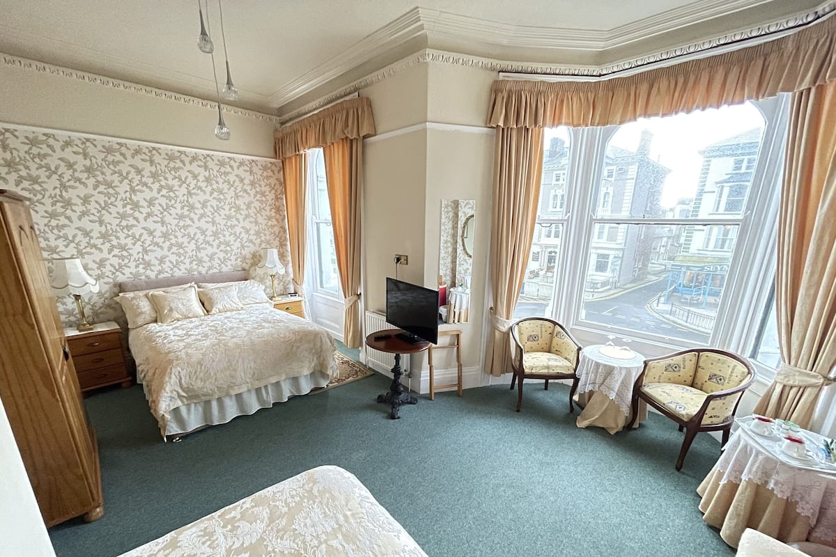 A hotel room with a large bed, a television, two armchairs, a small table, and a view of the city through large windows.
