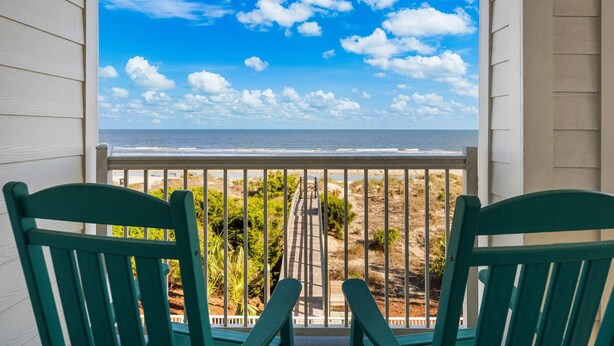 Ein Balkon mit zwei grünen Stühlen, einem Holzboden und Blick auf Strand und Meer.