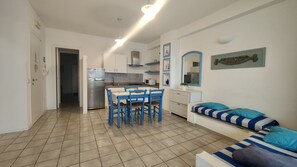 A compact kitchen with a refrigerator, microwave, and stove, a dining area with blue chairs, and a striped sofa with pillows.
