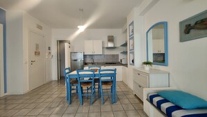 Cozinha compacta com armários brancos, forno embutido e pia. Há uma mesa de jantar com cadeiras azuis e um sofá branco com almofadas azuis.