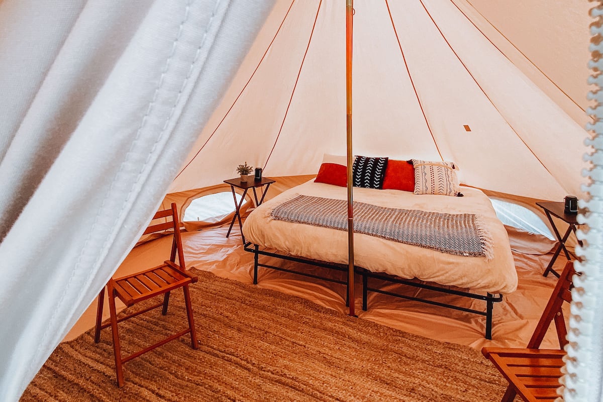 Een ruime tent met een bed, twee houten stoelen en een kleine tafel met een lamp.