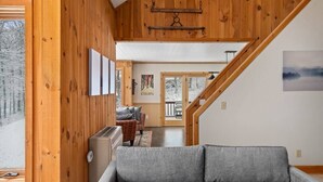 A cozy wooden interior with a grey sofa, wooden walls, a staircase, and a view of a snowy landscape. - Brand New Kitchen & Bathrooms, close to Mt Snow! (Dover)