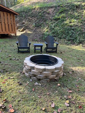 A stone fire pit with two Adirondack chairs and a small table in a grassy backyard. - Romantic Retreat Minutes from downtown Pigeon Forge! (Sevierville)